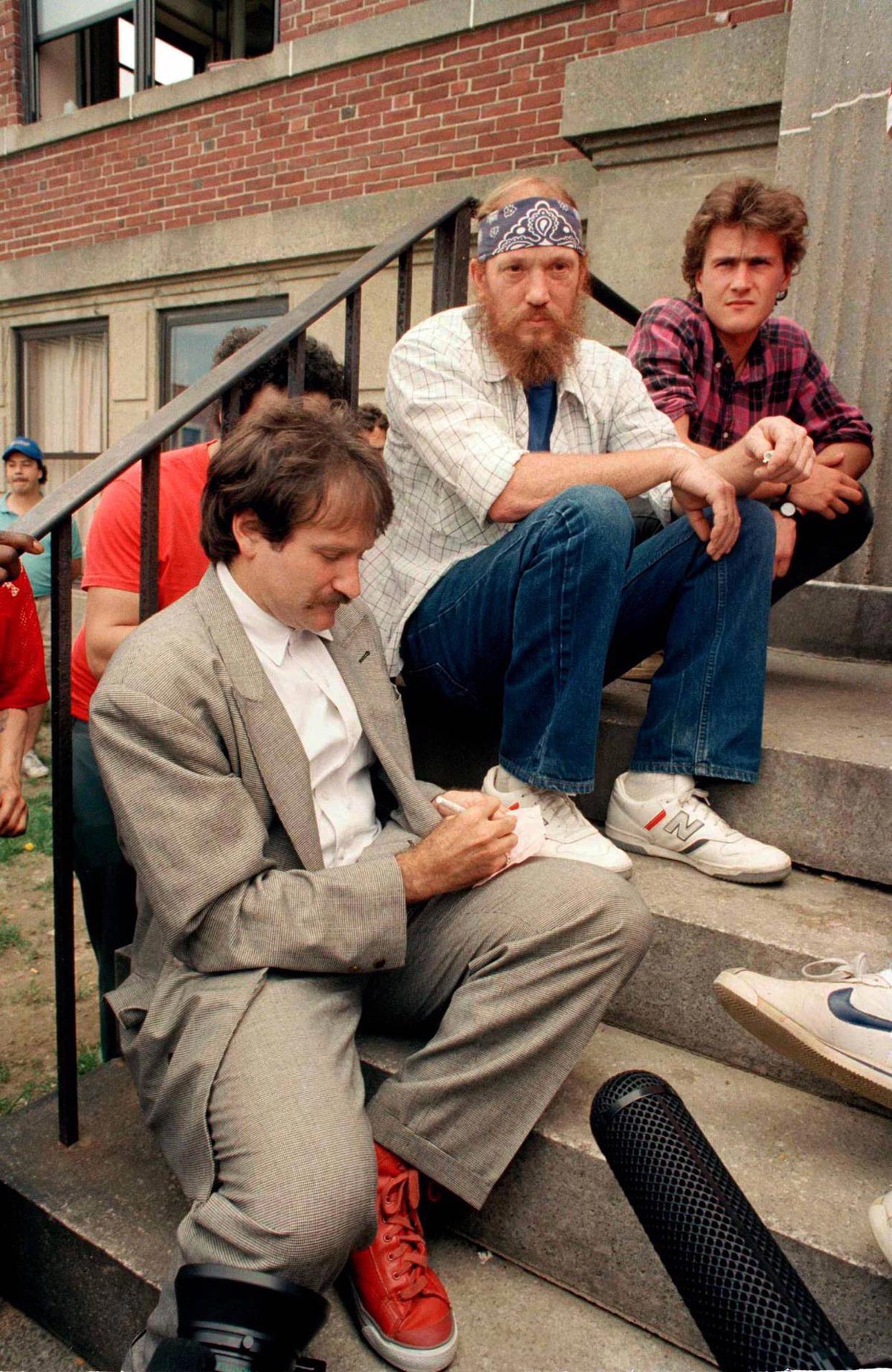 A man in a gray suit sits on outdoor steps, signing an autograph, while two casually dressed men with beards and plaid shirts sit on the steps behind him. A brick building and other people are visible in the background.