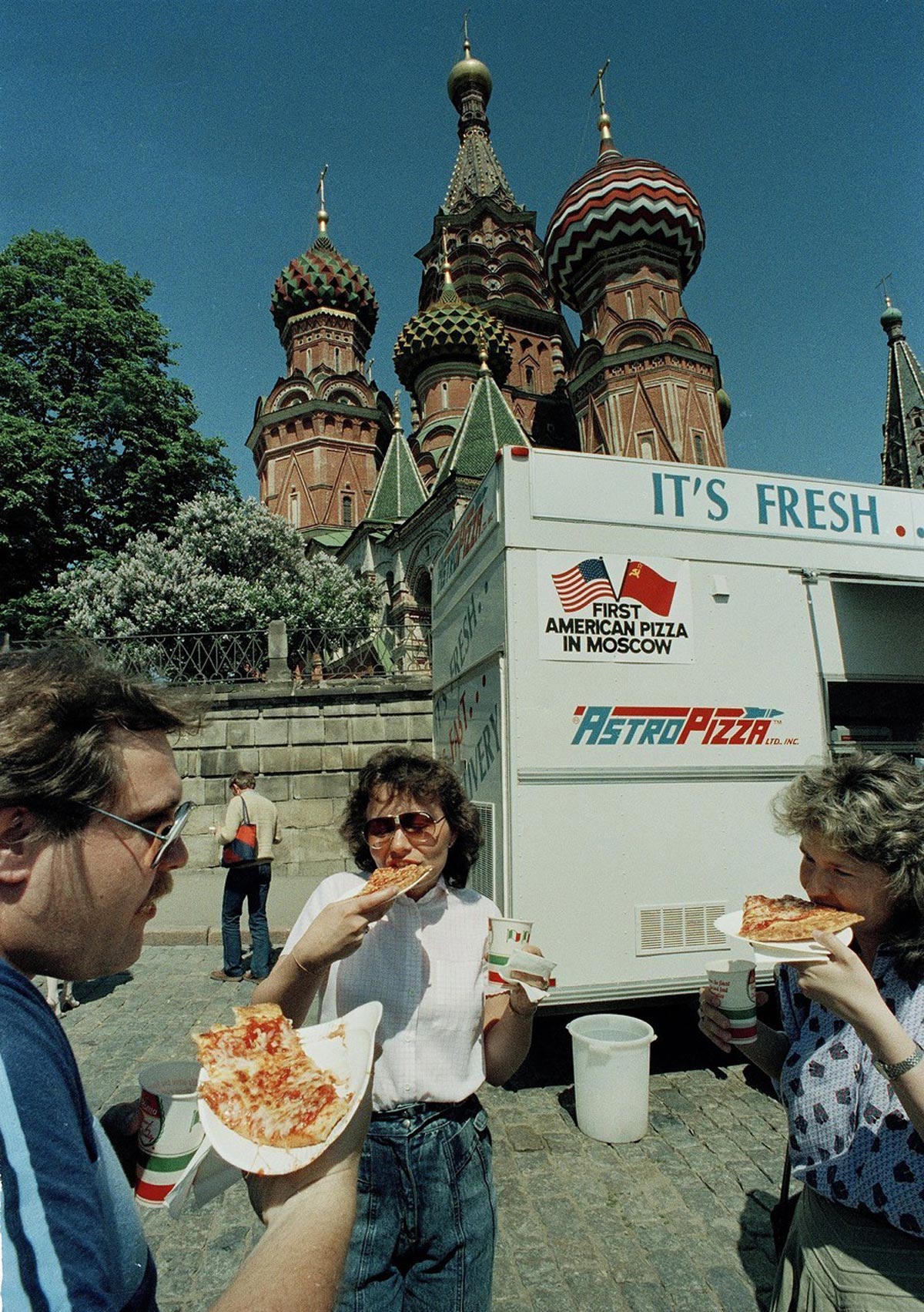 Three people eat pizza and drink soda in front of a food truck labeled First American Pizza in Moscow near Saint Basil’s Cathedral, with the cathedral’s colorful domes visible in the background.