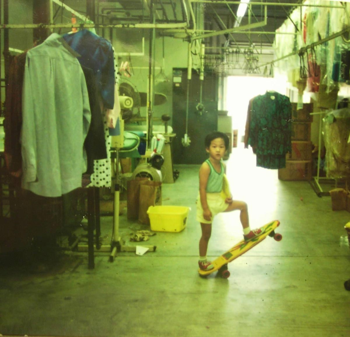 A young child stands with one foot on a skateboard inside a laundromat or dry cleaning shop, surrounded by hanging clothes and industrial laundry equipment. The scene is brightly lit and has a nostalgic feel.
