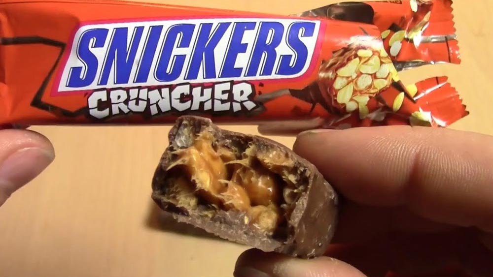 A hand holds a partially unwrapped Snickers Cruncher bar, revealing its chocolate-covered, caramel, and crunchy nut filling. The wrapper is visible in the background.