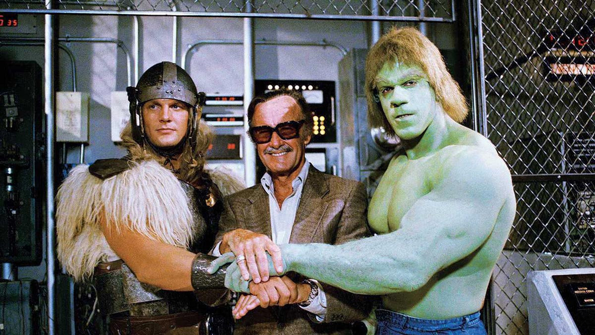 Three men stand together: one dressed as a Viking-like warrior, one wearing sunglasses and a suit, and one painted green with muscular arms, resembling the Hulk, all smiling and clasping hands in front of control panels.