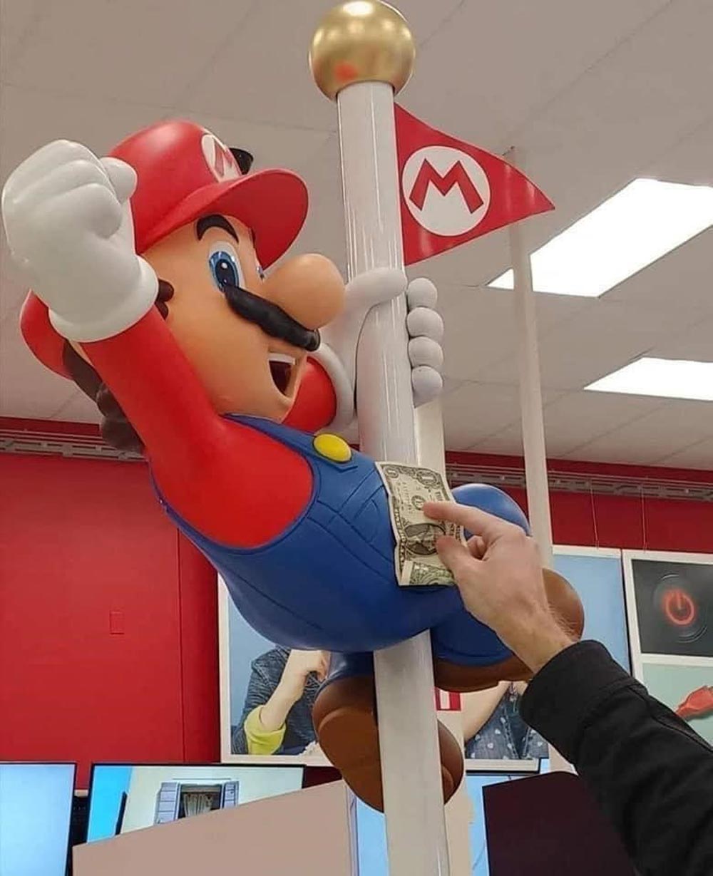 A large Mario statue holding a flagpole is in a store. A person’s hand is placing a dollar bill in Mario’s overalls, as if tipping him. Red and white Mario-themed decor is visible in the background.