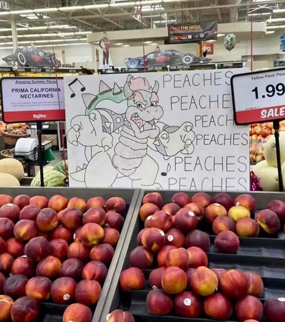 A grocery store display shows nectarines below a hand-drawn sign of Bowser from Mario, singing Peaches, peaches, peaches with musical notes. The fruit is mislabeled as nectarines instead of peaches.