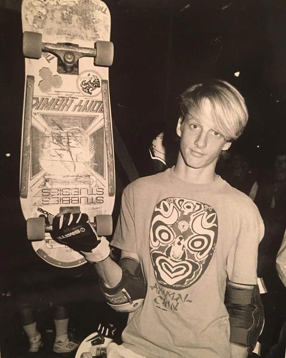 A teenage skateboarder with straight blond hair holds up a skateboard vertically, wearing a graphic t-shirt, gloves, and pads. The image is in black and white and taken indoors, with several people blurred in the background.